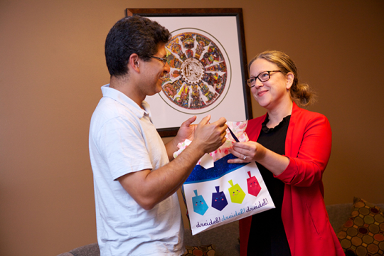 an image of two adults; a woman handing a plastic bag with pictures of dreidals and the word dreidal on it to a man