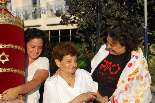 Three people standing while one of them holds a Torah with a red cover