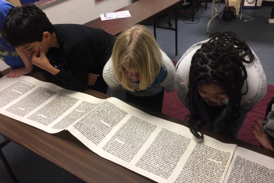 Three children reading a megillah placed on a brown table