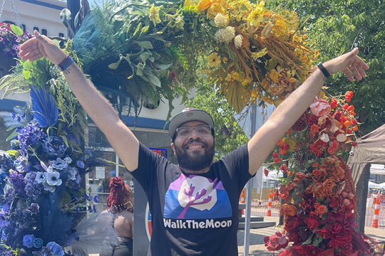 Photo of Chaim Harrison wearing a Walk the Moon shirt, holding his hands up, standing in fromt of a rainbow colored arch