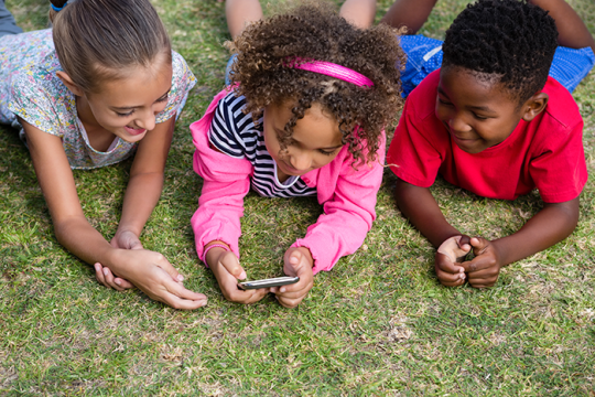 Photo of 3 kids using mobile phone while lying on field in yard