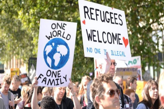 Protesters holding signs that read ONE WORLD ONE FAMILY and REFUGEES WELCOME NO ONE IS ILLEGAL