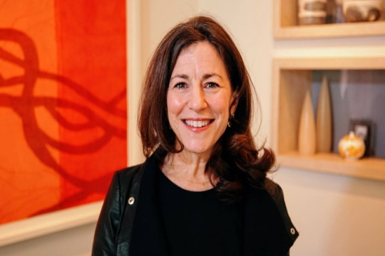 Headshot of Rabbi Carole Balin smiling at the camera against a blurred orange and red background