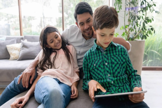 Dad and two smiling kids looking at an iPad together on the couch