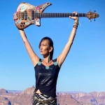 Benally looks to the left while raising a base guitar above her head in front of a clear blue sky
