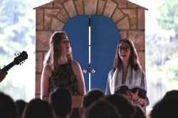 Two young women wearing prayer shawls stand at a bimah and lead a crowd of teens in worship