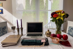 Home sanctuary setup of a table in front of a window bearing a laptop surrounded by flowers and Jewish ritual objects like a prayer book and candlesticks