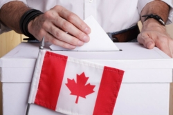 A mans hands dropping a ballot into a ballot box with a Canadian flag in front