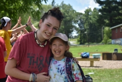 Smiling camp counselor with her arm around a smiling young camper in a hat 