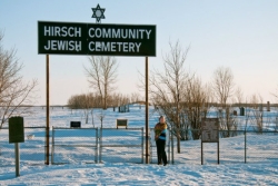 A smiling woman in the snow in front of a large sign reading HIRSCH COMMUNITY JEWISH CEMETERY