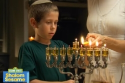 Screenshot of Shalom Sesame video video of young boy in a kippah watching an adult light an oil hannukiyah