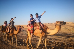 Teens smiling and waving while riding camels in the desert