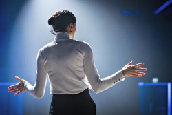 Woman in a white turtleneck with her back to the camera and her hands up as if speaking to an audience 