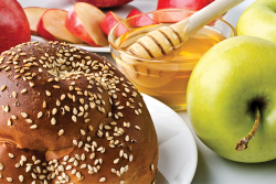 A table with sliced apples, a bowl of honey with a wand, and a round challah.