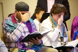 an image of two people sitting down, wearing yamukas and tallit, covering their eyes and praying, with prayer books in hand