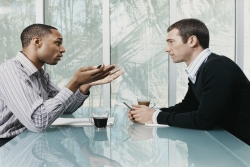 Photo of two men talking, sitting at a table with coffee in front of them
