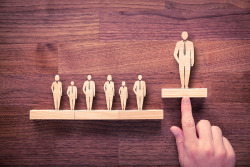 Photo of multiple wooden people figurines on two shelves; one figurine is on a shelf higher then the rest