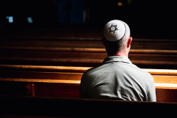Photo of a man sitting in an empty, dark sanctuary wearing a white yamuka with a blue star of david on it