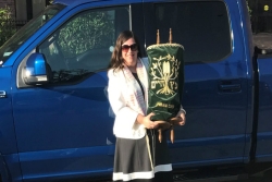 Female rabbi in a black and white dress holding a Torah scroll while standing in front of a blue truck 