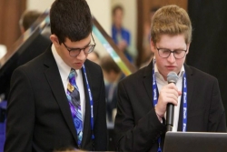 Two teen boys in suits reading from a document in front of a crowd of other teens 