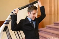 Smiling bar mitzvah boy wearing a suit and kippah and wrapping himself in a prayer shawl from the bimah