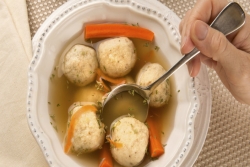 Closeup of a hand dipping a spoon into a full bowl of matzah ball soup with carrots