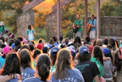 Image taken from the back of an outdoor sanctuary space with a rabbi on the bimah at the front and showing the backs of the heads of campers with their arms around one another