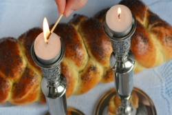 Hand lighting Shabbat candles in silver candleholders next to a challah