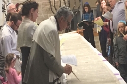Man using a yad to read from an unfurled Torah scroll on a table as members look on