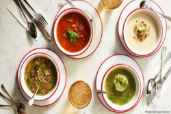 Overhead photo of four different soups in bowls with silverware, napkins, and drinks around the bowls
