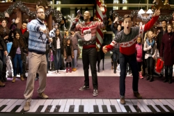 Men in Hanukkah and Christmas sweaters dance on a lifesized piano