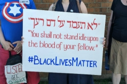 White womans hands holding a sign in Hebrew and English reading YOU SHALL NOT STAND IDLE UPON THE BLOOD OF YOUR FELLOW and concluding with the phrase BLACK LIVES MATTER