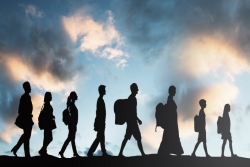 Eight individuals (adults and children) in shadow walking single file in front of a sky of sun and clouds