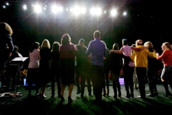 Group of people with their arms around one another standing on stage and facing an audience 