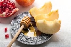 Apple slices and a honey stirrer on a silver platter carved with scenes of Jerusalem and a pomegranate in the background