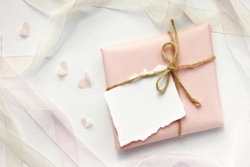Small flat gift wrapped in light pink paper and decorated with a homemade bow and handmade card; pink and white ribbons and small cut-out hearts are in the background