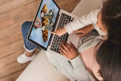 Little girl sitting on her mothers lap and pointing at a laptop screen where a hand is lighting shabbat candles next to a loaf of challah