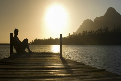 Person in shadow sitting on the edge of a dock; arms and hands around knees