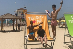 Three men in bathing suits sitting on a large chair on the beach that reads TEL AVIV LOVES ME