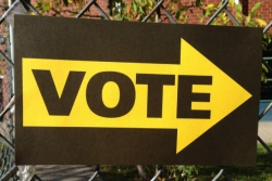 Large VOTE sign hanging on a chain link fence with an arrow pointing toward polling place