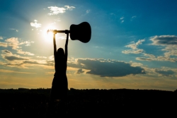 Woman in shadow facing away and holding a guitar overhead with both hands. Sunshine and clouds surround her shadow.