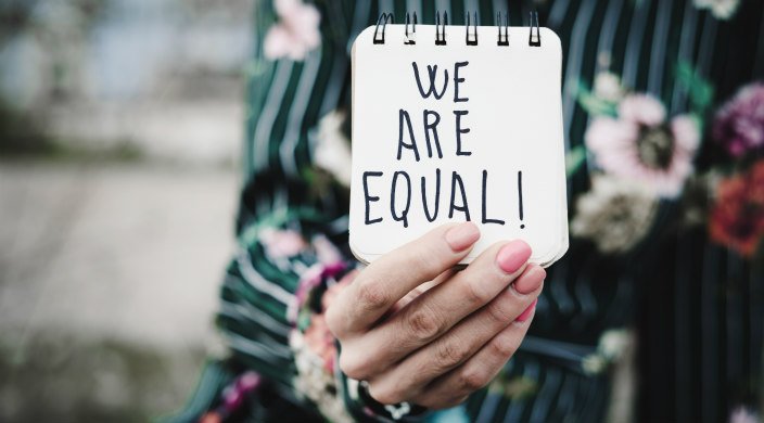 Woman (from the neck down) holding small notepad that says: "We Are Equal!"