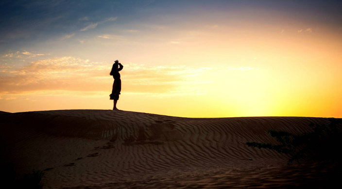 Silhouette of a woman standing alone on a desert sand dune against a sunset